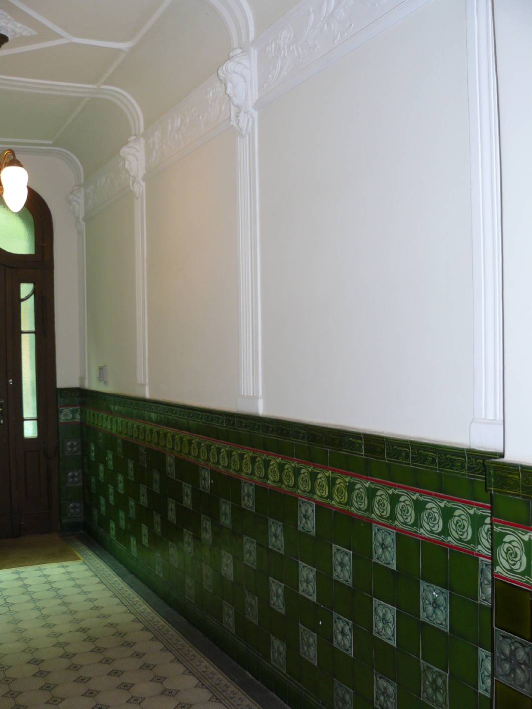 Im Eingangsbereich Esmarchstraße Berlin wurde vom Maler – Innungsbetrieb und Malermeister & staatl. geprüfter Techniker Norbert Schimpf die Fliesen aus Stuck – Gips nachgeformt und mit einer Schablonierung in Lasurtechnik bemalt. Der Stuck wurde restauriert (Restaurator). Als Treppenhausgestaltung wurde in dem Jugendstil – Treppenhaus eine Holzmalerei und eine Marmormalerei in Form einer dezenten Marmorierung als  Lasur und Kalkspachtel – Glättetechnik angebracht. Die um das Treppenhaus leicht zu gestalten wurde eine helle Marmorimitation gewählt. Zuvor wurden in dem Altbau – Treppenhaus und Entree die Malerarbeiten sorgfältig ausgeführt. An den Wangen hat der Malerbetrieb Restaurierung & Gestaltung / Exklusive Malerarbeiten Stuck angeracht und mit einer Holzimitation bemalt. Die Traljen wurden mit Schelllack – Lasur als Holzimitation gestrichen. An den Stufen und Türflächen wurde eine Holzlasur mit Holzmaserung in einer Holzmalerei als Lasurtechnik abmalt. An den Decken wurde eine Deckenmalerei mit Eck Schablone und Begleitstrich als Schablonentechnik schabloniert. Das Ornament der Deckenmalerei wurde von den Türornamenten abgenommen. Die zum Teil aus Stuck – Gips nachgeformt und mit einer Holzimitation bemalt wurden.